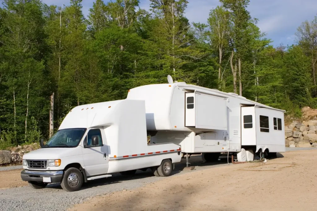White fifth-wheel trailer coupled to a truck on a gravel campsite pad in the trees