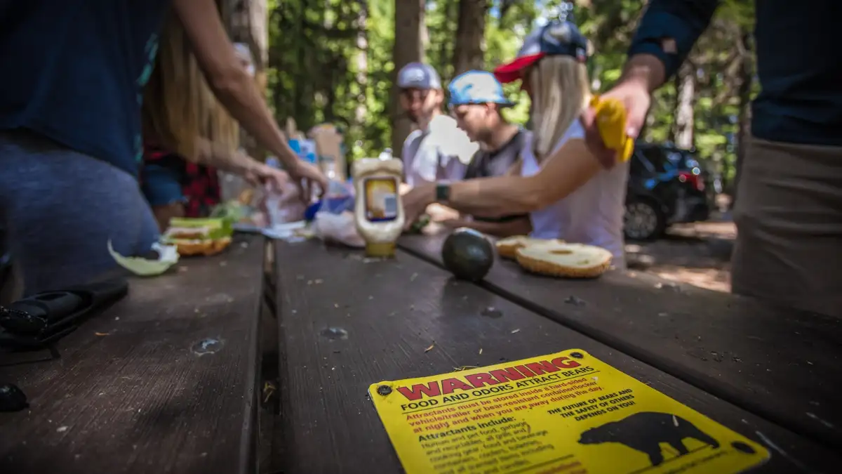 Campers preparing a meal at a Glacier National Park campground with a bear safety sign in the foreground.