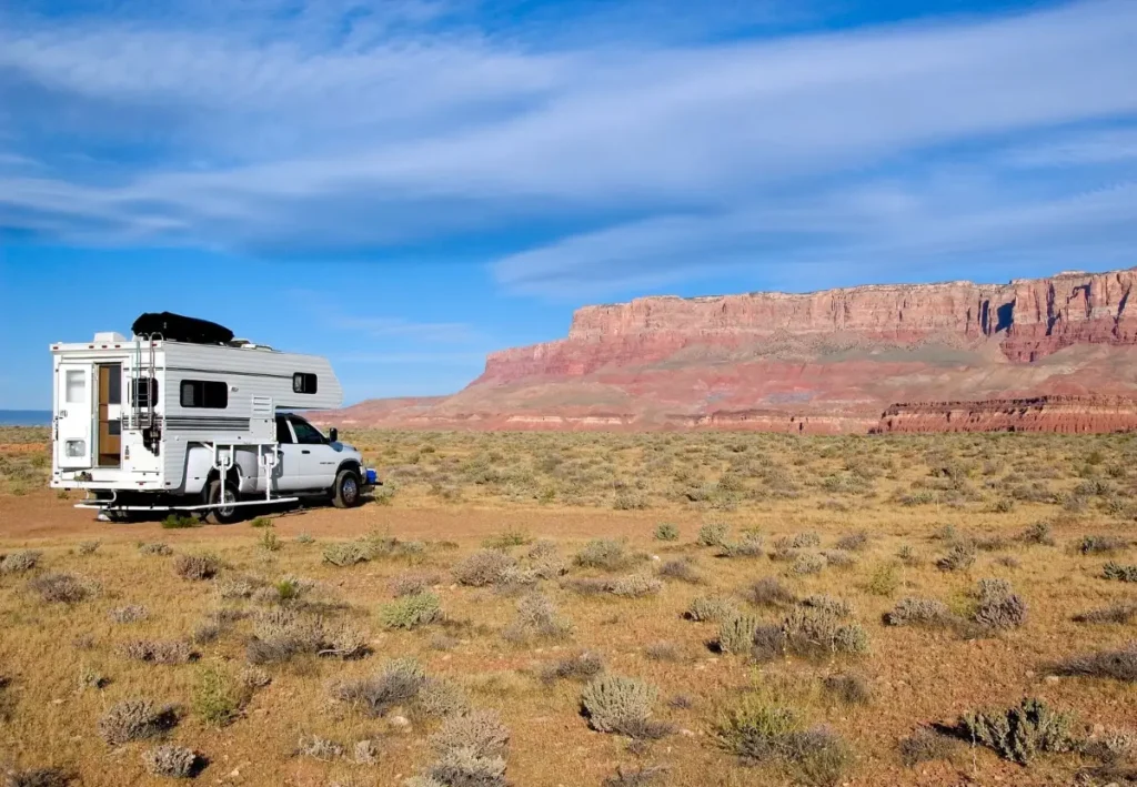 Truck camper parked alone on open desert scrub with red cliffs behind